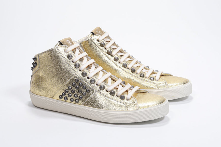 Three quarter front view of mid top metallic gold sneaker. Full leather upper with studs, an internal zip and vintage rubber sole.