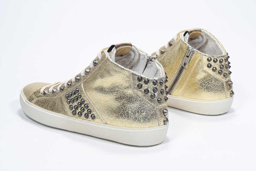 Three quarter back view of mid top metallic gold sneaker. Full leather upper with studs, an internal zip and vintage rubber sole.