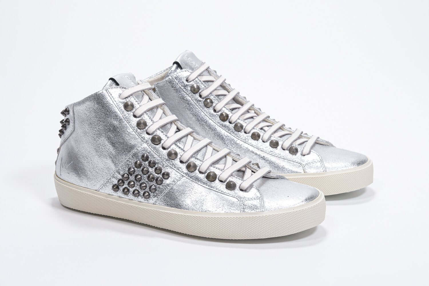 Three quarter front view of mid top metallic silver sneaker. Full leather upper with studs, an internal zip and vintage rubber sole.
