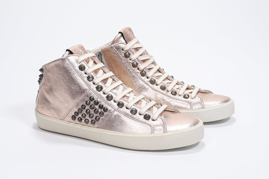 Three quarter front view of mid top metallic rose sneaker. Full leather upper with studs, an internal zip and vintage rubber sole.