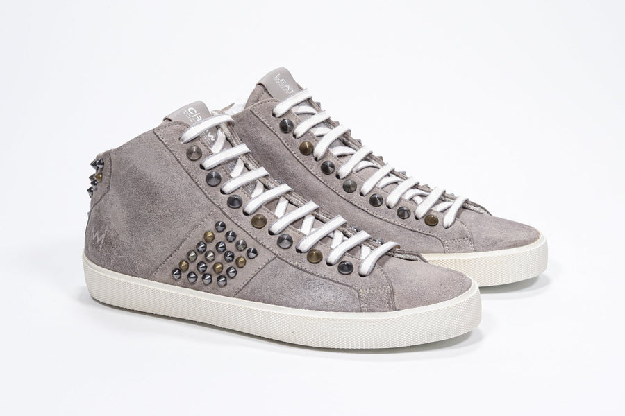 Three quarter front view of mid top beige color sneaker. Full suede upper with studs, an internal zip and vintage rubber sole.