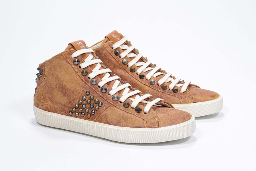Three quarter front view of mid top rust color sneaker. Full suede upper with studs, an internal zip and vintage rubber sole.