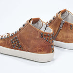 Three quarter back view of mid top rust color sneaker. Full suede upper with studs, an internal zip and vintage rubber sole.
